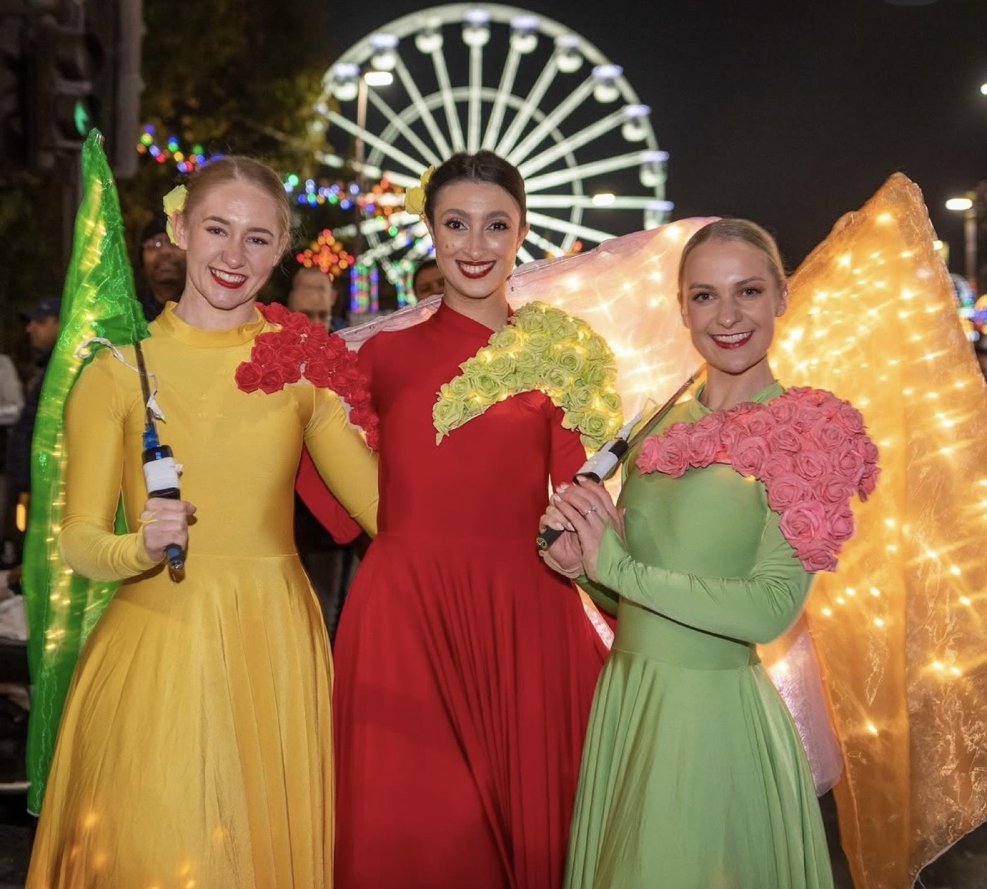 Three performers in illuminated yellow, red and green costumes with light-up wings at the Diwali Festival.