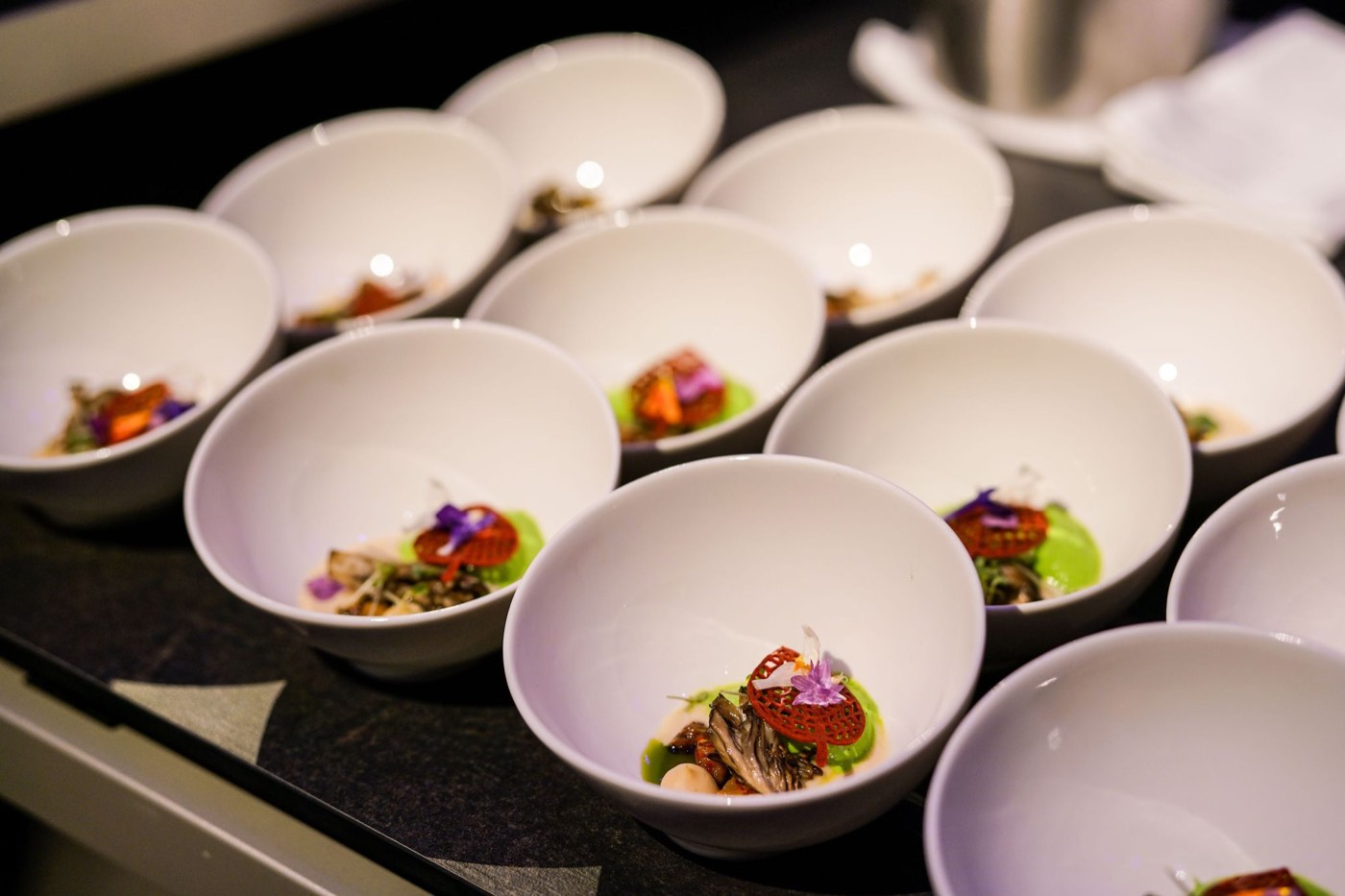 Rows of individual tasting bowls plated with mushrooms, herb purée and an edible flower, ready for service.