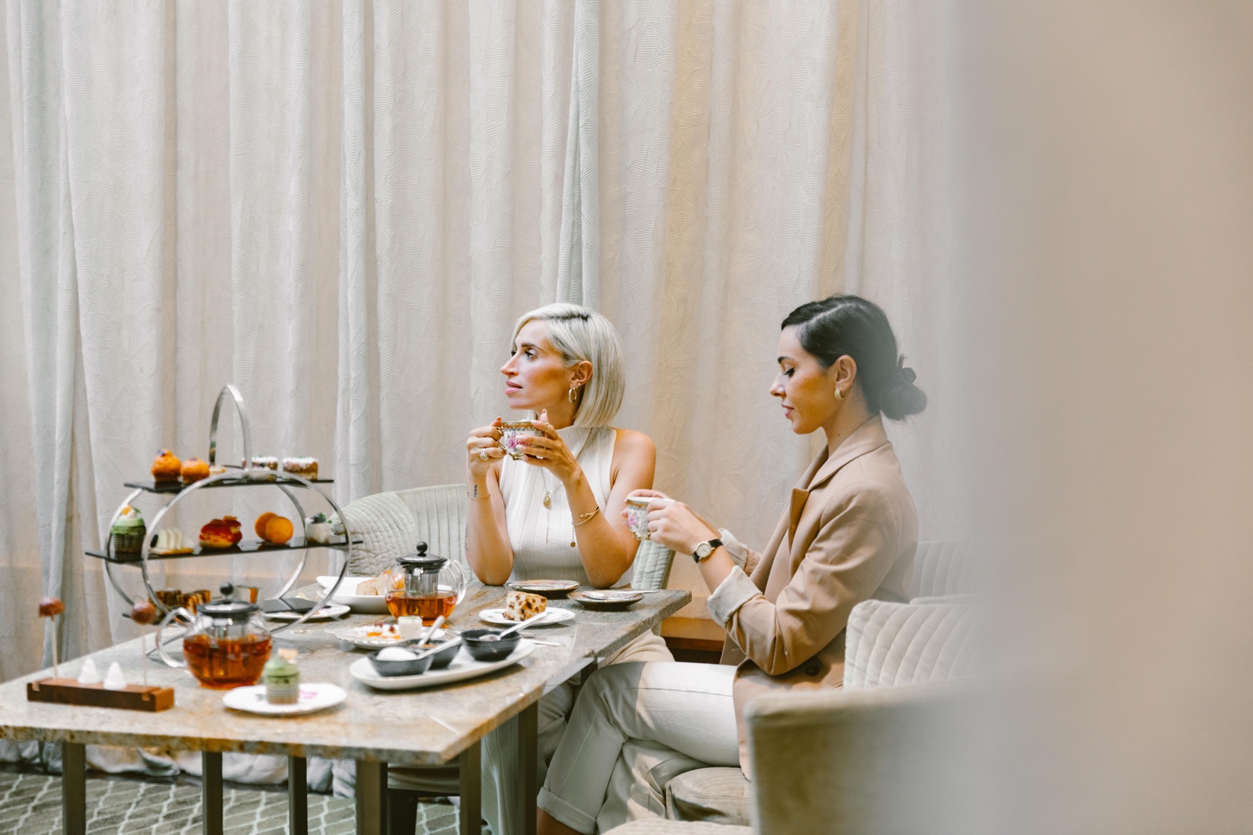 Two guests taking afternoon tea at Park Hyatt Zurich, teacups in hand beside a tiered pastry stand and marble table.