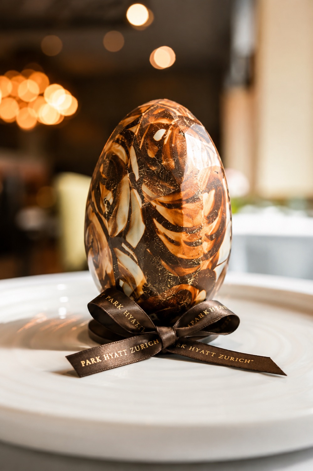 A marbled chocolate Easter egg finished with a brown Park Hyatt Zurich ribbon, plated under warm bokeh light.