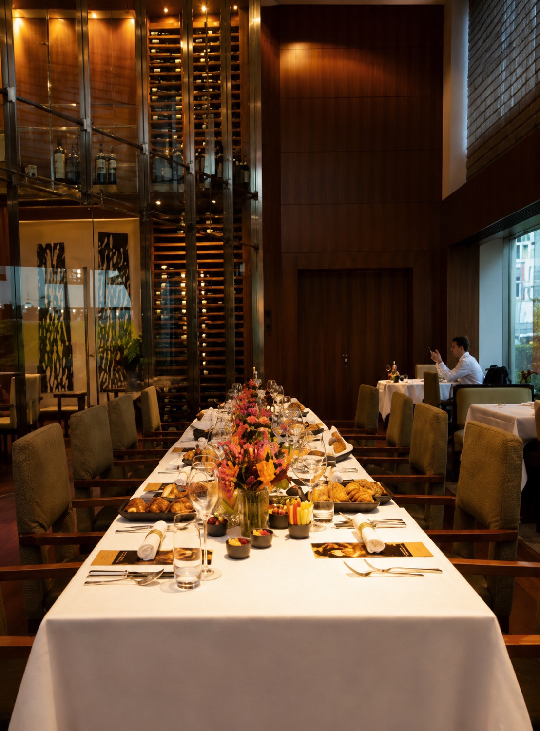 A long banquet table dressed in white linen at Park Hyatt Zurich, set for breakfast with pastries and a vivid floral centrepiece, framed by a towering glass wine wall.