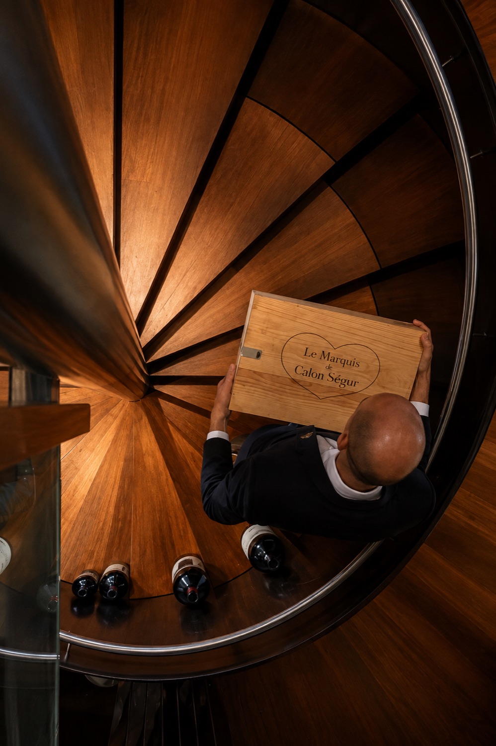 Overhead view of a sommelier descending the hotel's spiral wooden cellar staircase, carrying a wooden wine case past bottles resting on each tread.