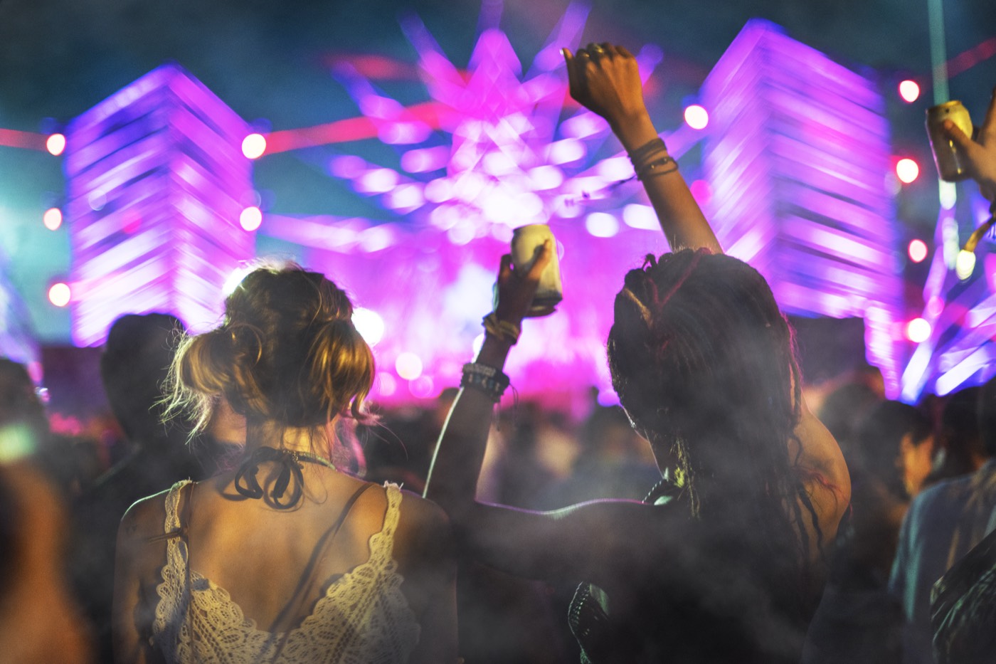A festival crowd at night with hands raised, silhouetted against bright stage lights.