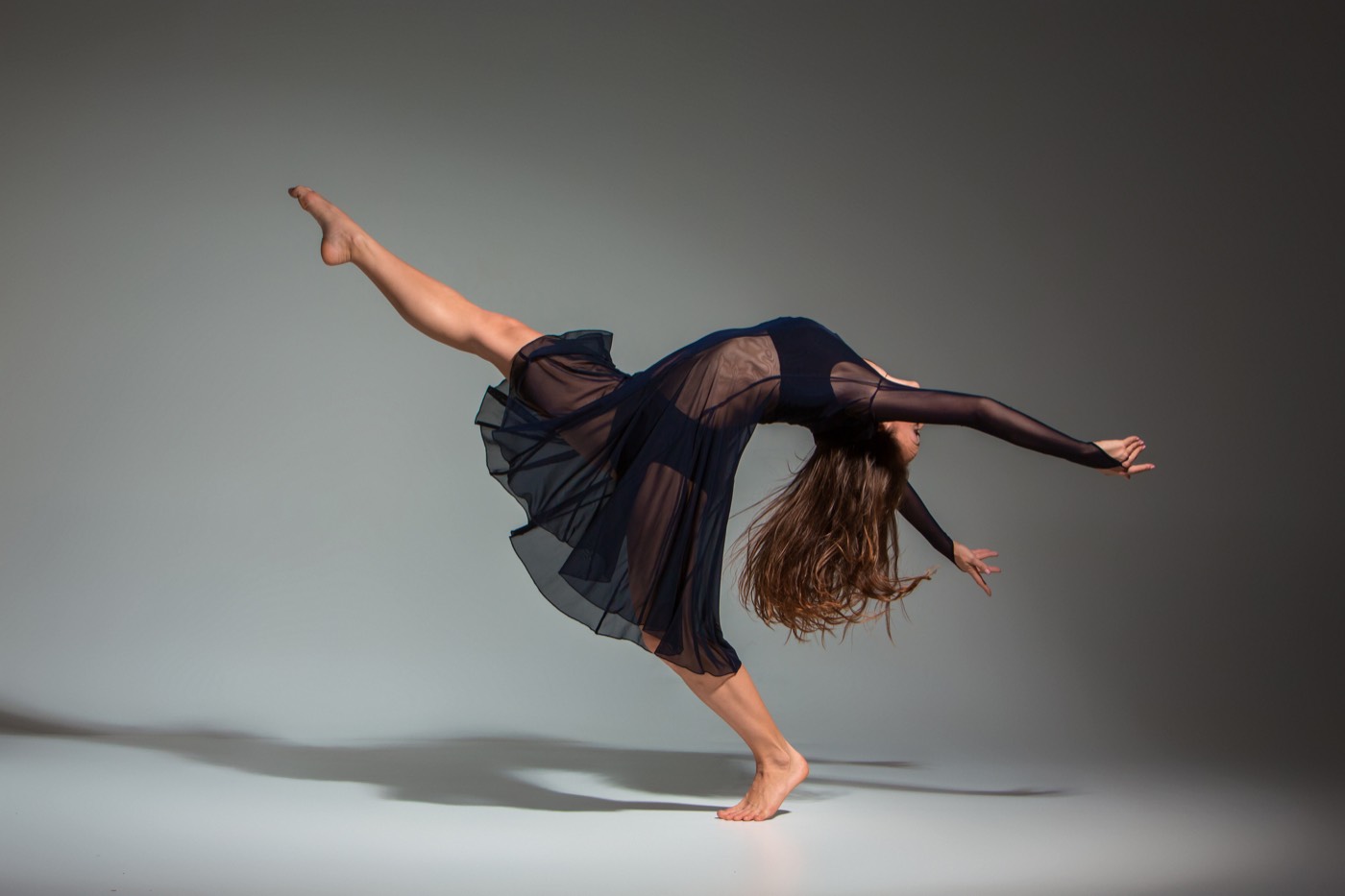 A contemporary dancer in a flowing dark dress arched mid-movement in a softly lit studio.