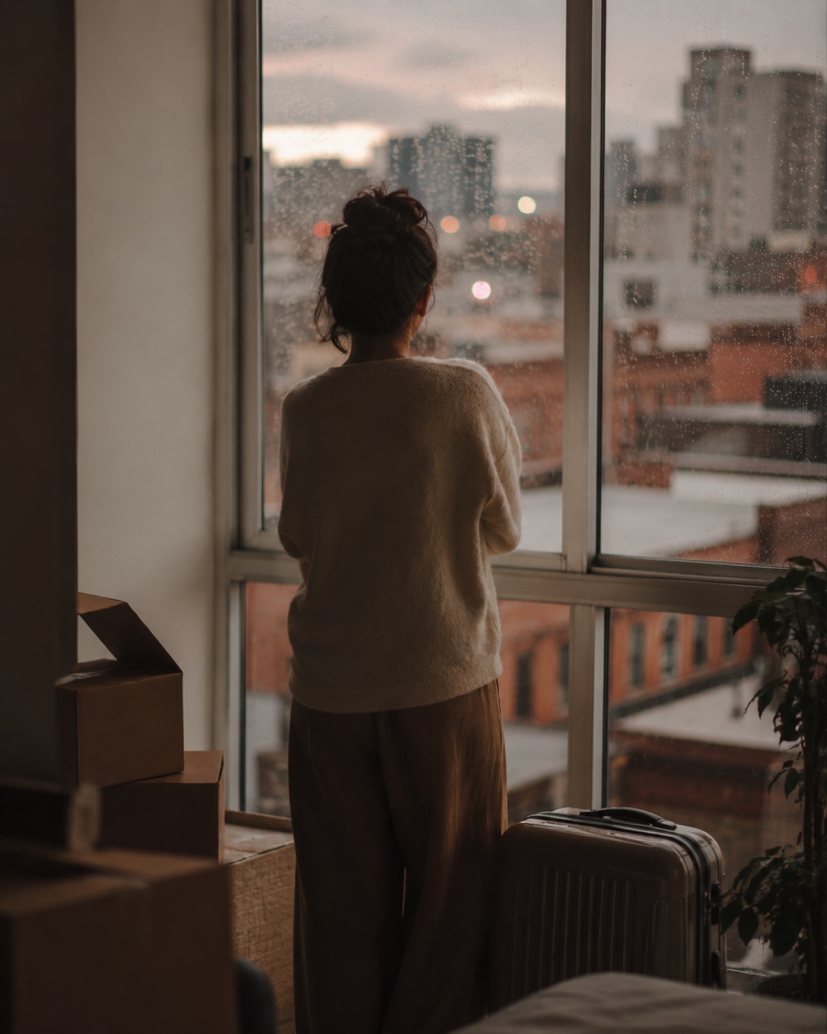A person seen from behind pausing at a rain-flecked window, city light soft beyond the glass — the quiet weight of rebuilding a life in a new place.