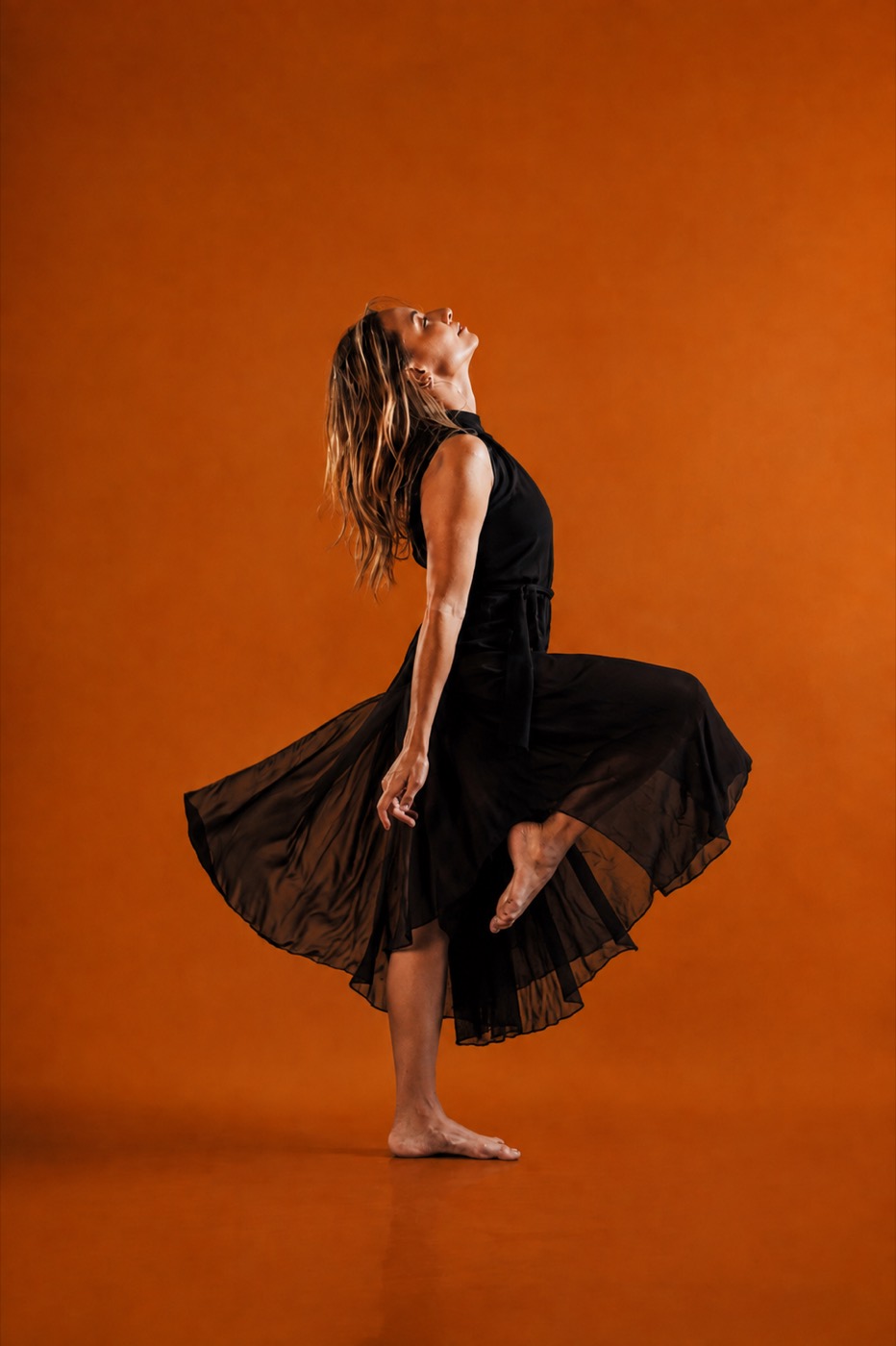 Luisa Sandoval in a black dress, back arched and hair swept back, against a warm orange backdrop.