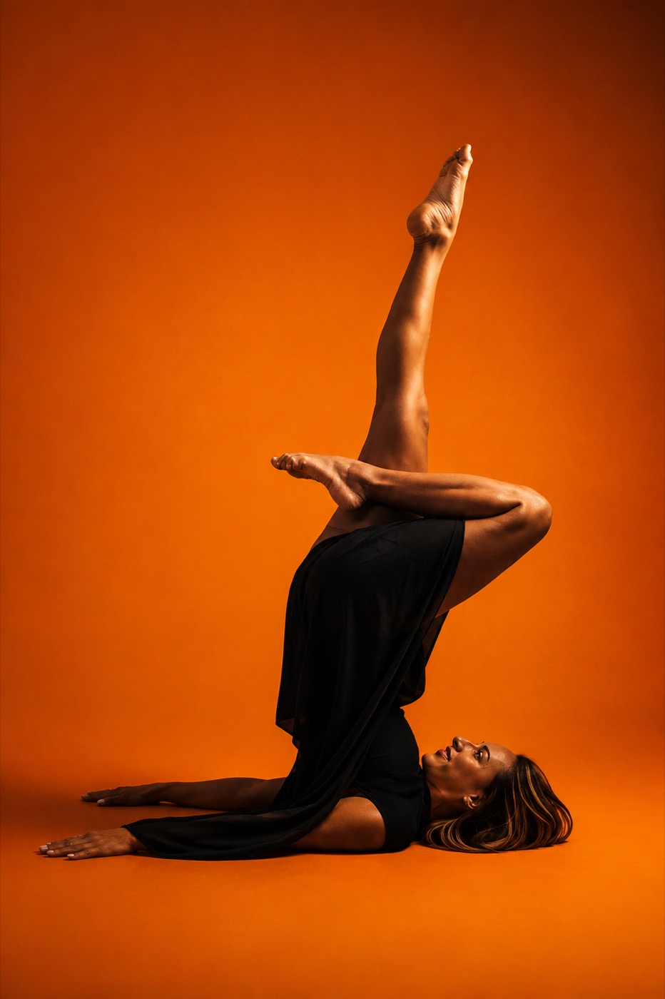 Luisa on the floor with one leg extended vertically, black fabric draping, against a warm orange backdrop.
