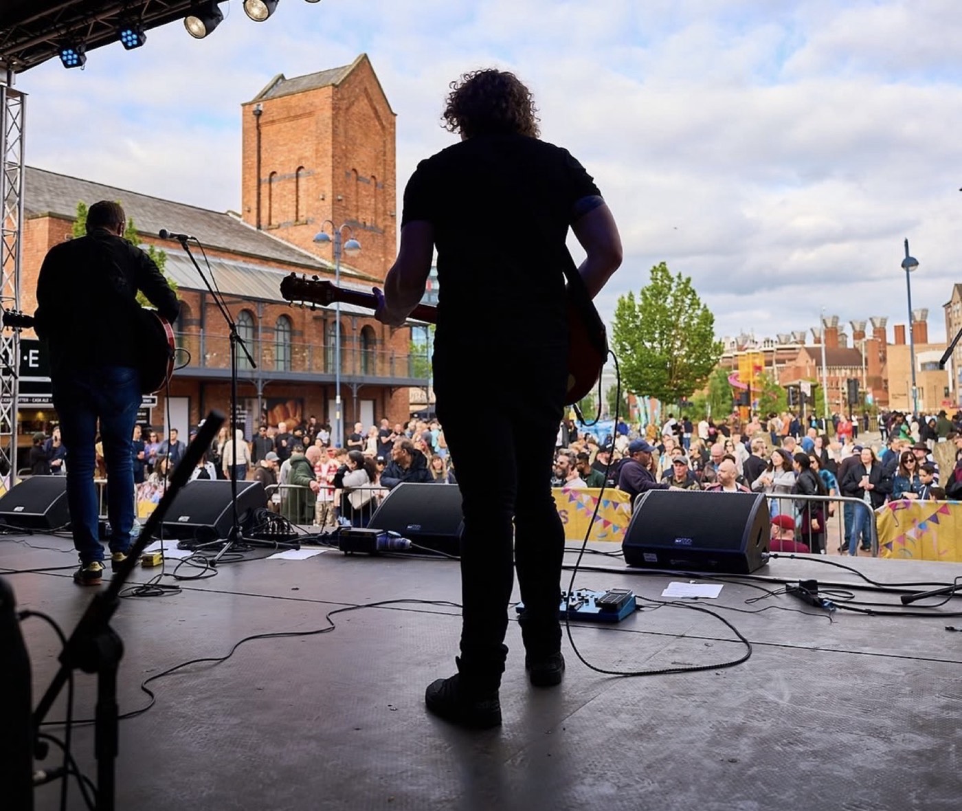 A live band performing on the Riverside Festival stage, viewed from behind the guitarist looking out over the audience.
